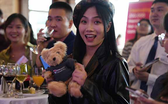 A member of the china alumni network smiling at the camera whilst holding a teddy bear with a Newcastle University jumper on